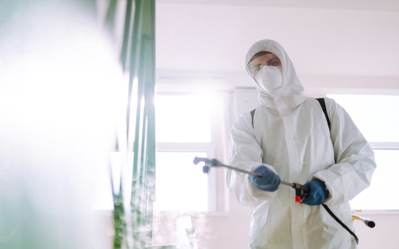 Man in Hazmat suit, protective gloves and goggles use sprayer equipment disinfect the entrance.